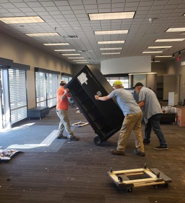The scene shows professional commercial junk removal: techs working together, focused on safety and control as they move the safe from inside the old Verizon office to the truck for proper disposal.