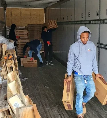 giant metal trailer-style storage unit. the doors are wide open, revealing it packed with crates, cardboard boxes, and miscellaneous household items. The technician, in a GoGo Junk Removal uniform, is carrying a large box out of the trailer while other crates and boxes are stacked near the opening, ready to be moved.