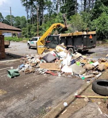 A large commercial property lot is being cleaned out after multiple incidents of illegal dumping. The ground is covered with scattered debris: broken furniture, construction waste, trash bags, pallets, and miscellaneous junk piled across the dirt and gravel.