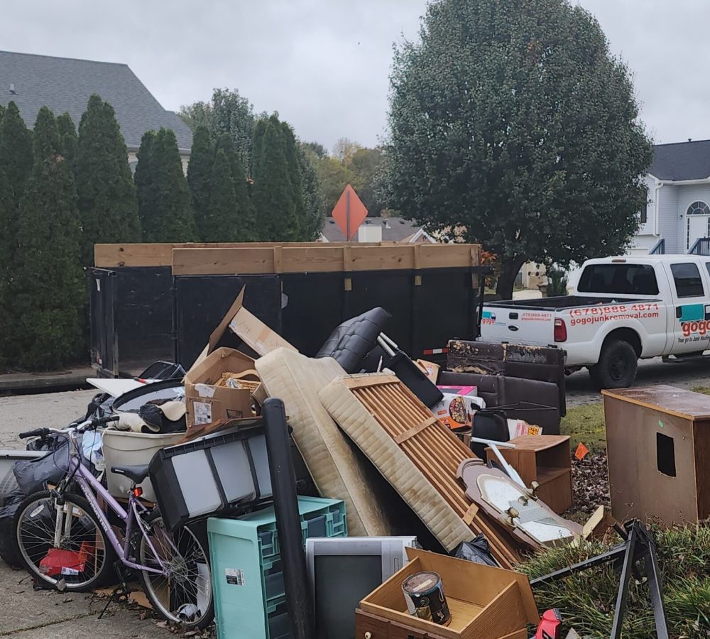 suburban curbside pickup in an Atlanta neighborhood. Along the curb there is a big pile of household debris and junk: old mattresses, a stove, a refrigerator, worn-out couches, boxes, and assorted household items stacked on the grass by the street.