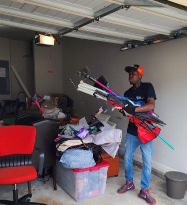A GoGo Junk Removal technician is working inside a residential garage, performing a garage cleanout. The garage door is open, and the tech in a GoGo uniform is carrying a mix of sorted debris—boxes, small household items, and light junk—out toward the driveway.