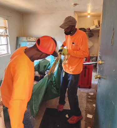 The scene captures professional, indoor apartment junk removal in progress, with the techs carefully hauling the green debris bag out of the building to take it to their truck.