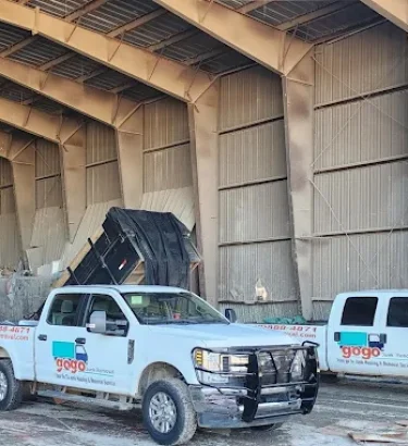 Two white Ford F-250 GoGo Junk Removal trucks are parked side by side at a transfer station. Each truck is hitched to a bumper-pull dump trailer. Both trailers are raised up in the air, actively dumping loads of mixed junk and debris onto the designated tipping area.