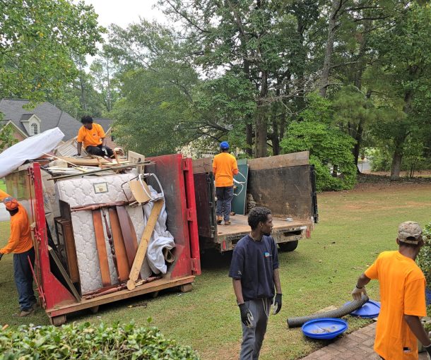 Several GoGo Junk Removal technicians are working a large estate/foreclosure cleanout at a residential property. Six or seven techs in matching uniforms are moving back and forth between the house and a row of large 40-yard dumpsters parked outside.