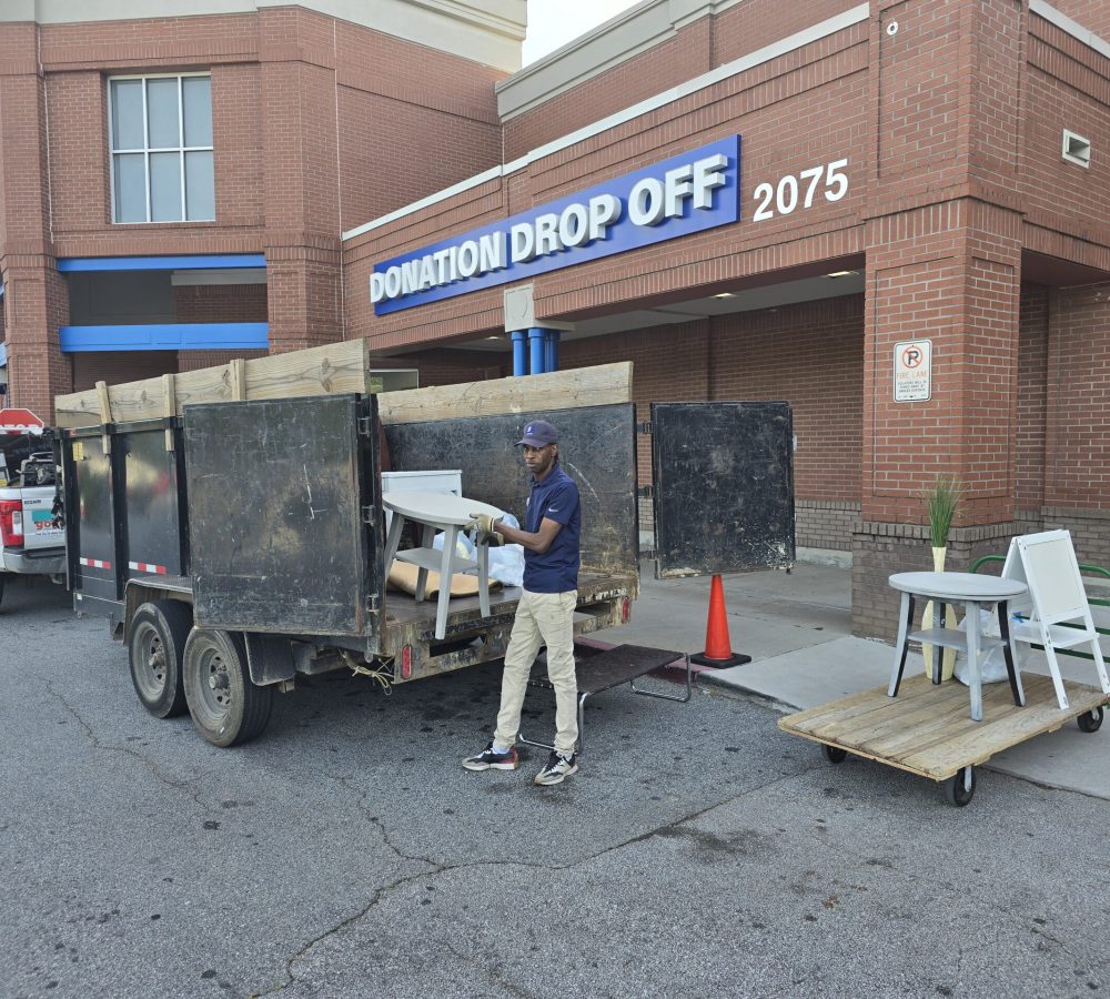 A truck and bumper pull trailer are at the donation center. A worker from the center is unloading donated items from the back of the trailer